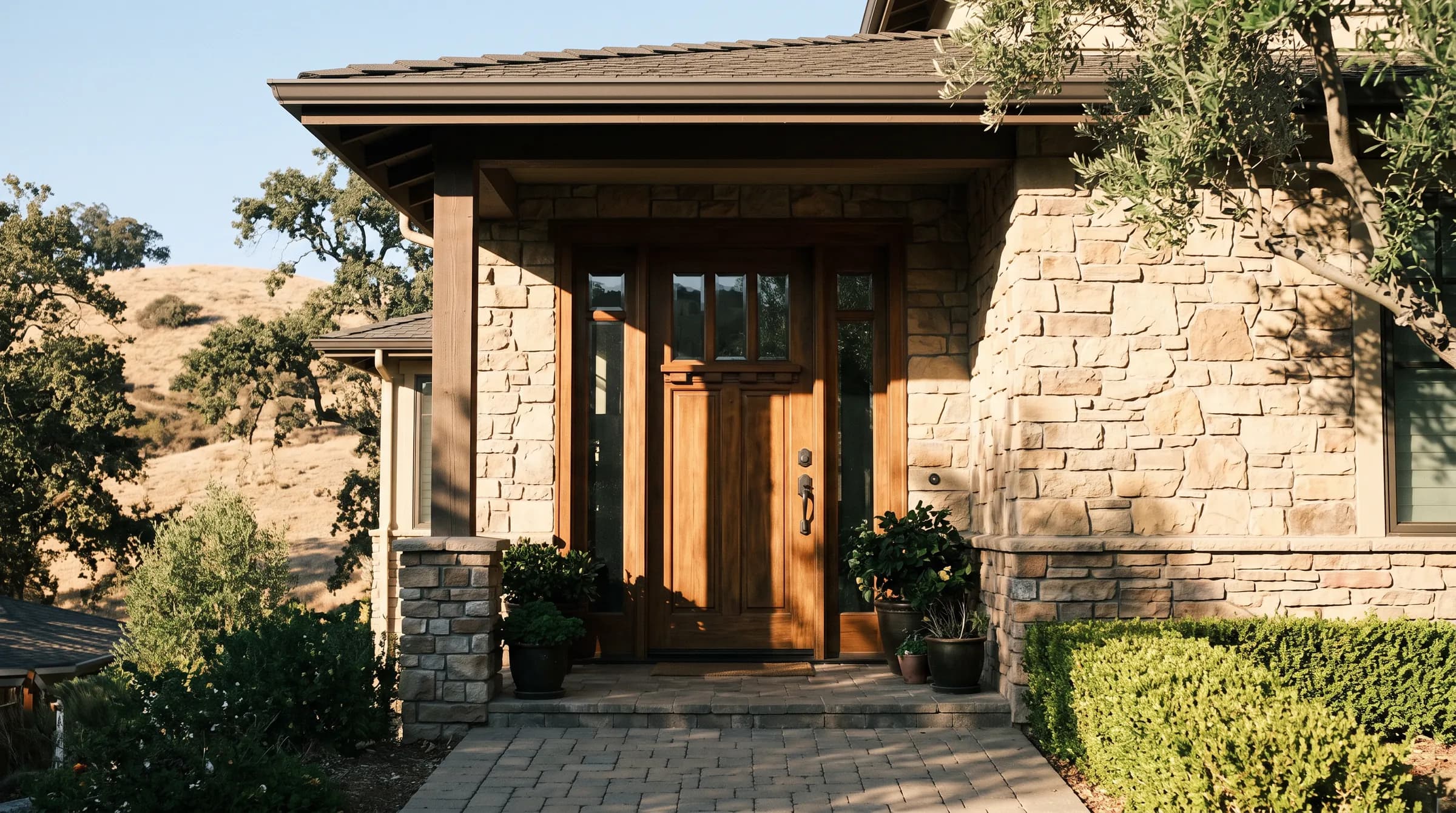 A Danville entry in the late afternoon: a wide painted door with a narrow glass sidelight beside it, a bronze handleset at hand height, the strike plate on the jamb catching the last sun, and the dry oak hills standing behind the roofline.